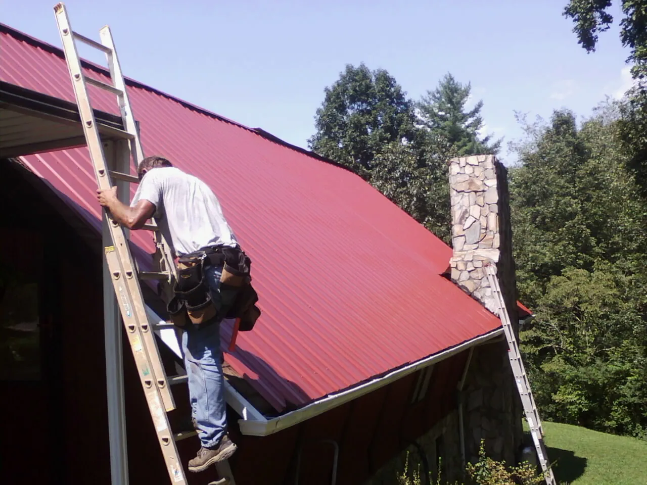 Roofer on a ladder beside a red metal roof and stone chimney — Soffit & Fascia Repair by Roofing Brothers in Fidelity